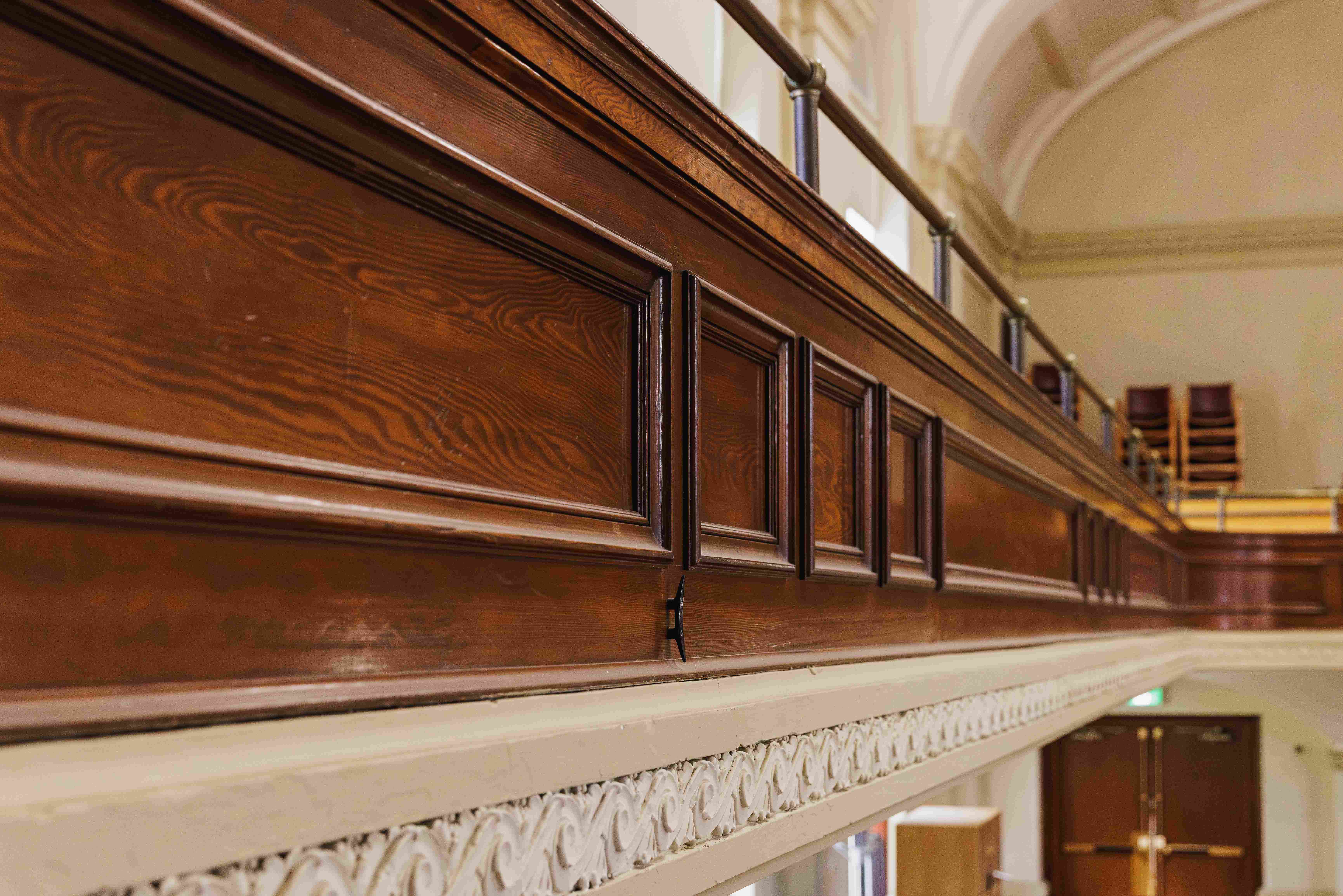 Close-up of historic wood-paneled balcony detailing with rich grain patterns, decorative molding, ornate trim, and architectural craftsmanship.